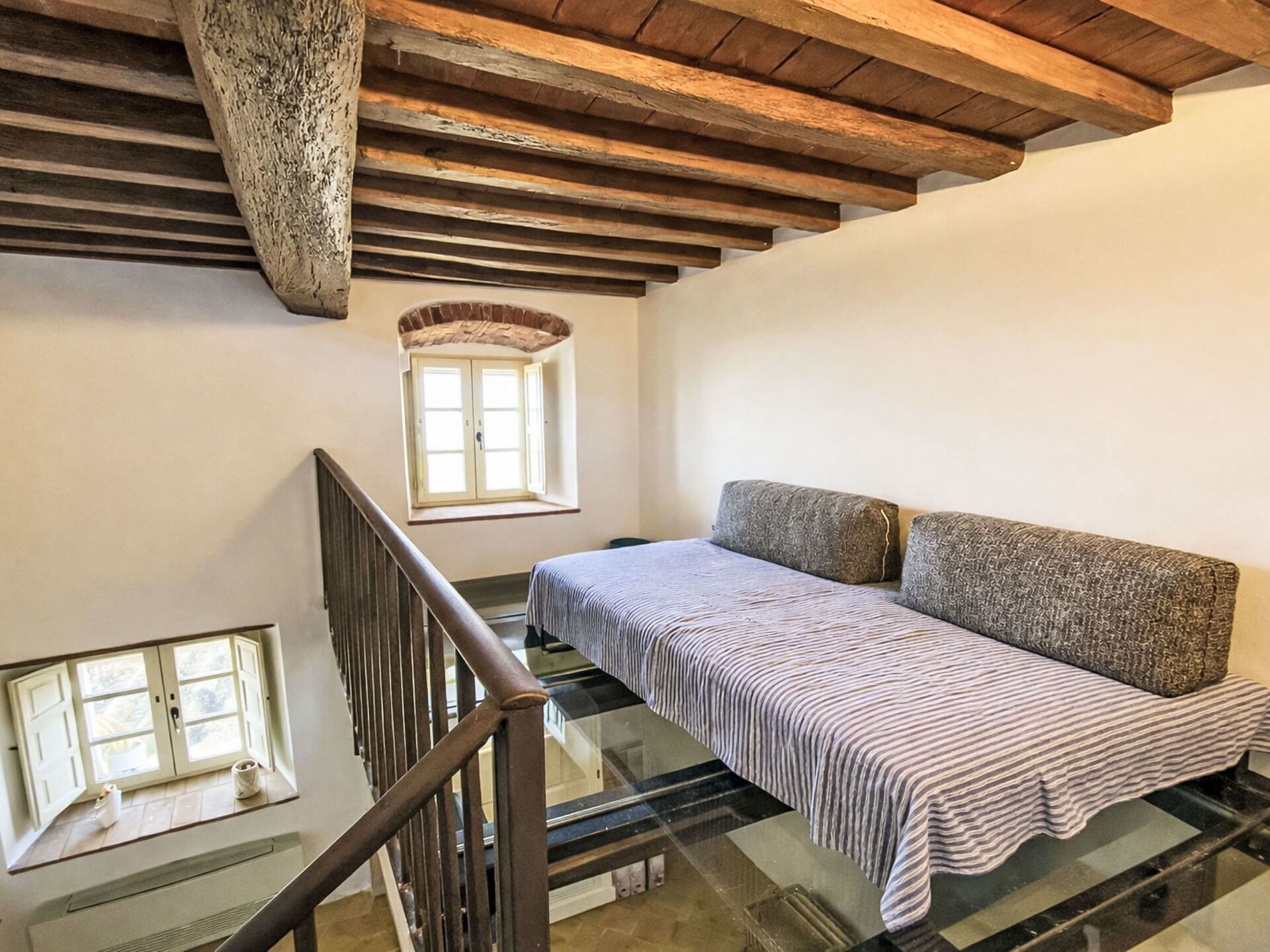 A room with a glass staircase, a bed with a striped bedspread, a sofa with patterned cushions, and a window with a view.