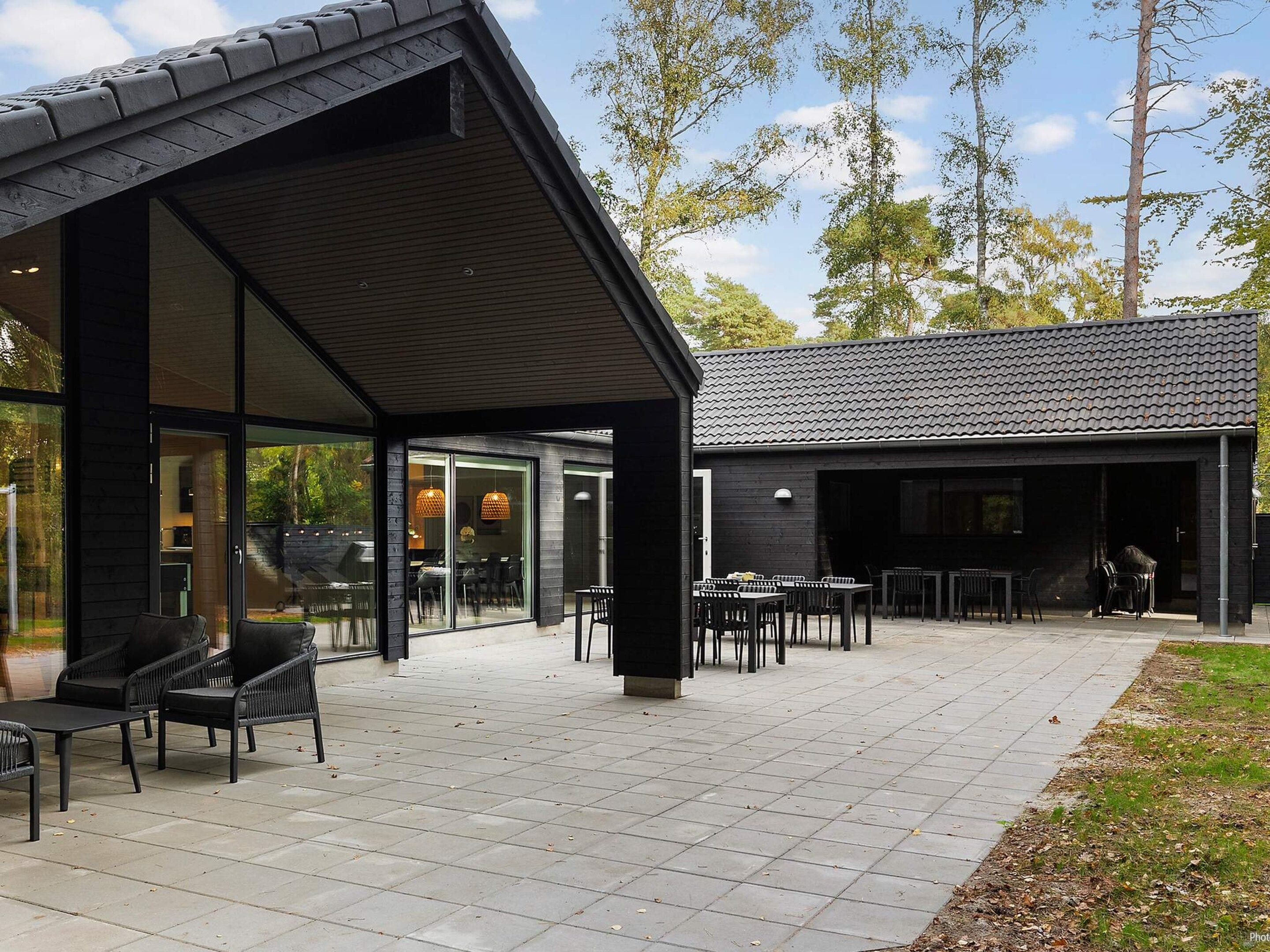 A modern outdoor patio area with a dining table and chairs, a covered seating area, and a building with a dark exterior and tiled roof.