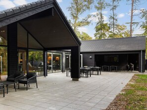 Ein moderner Außenbereich mit Terrasse, einem Esstisch und Stühlen, einer überdachten Sitzgruppe und einem Gebäude mit dunkler Fassade und Ziegeldach.