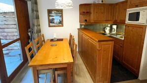 Een houten eettafel met stoelen, een keuken met houten kastjes, een magnetron en een hangende hanglamp.