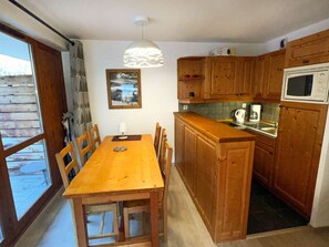 A wooden dining table with chairs, a kitchen with wooden cabinets, a microwave, and a hanging pendant light. - Cozy apartment - 6 guests (Valloire)