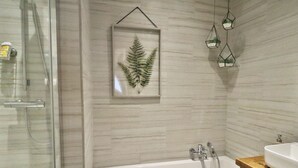 A bathroom with a glass shower enclosure, a framed fern artwork, a white bathtub, and hanging glass containers.