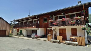 A two-story wooden building with a balcony, a car parked in front, and a wooden table and chairs outside.