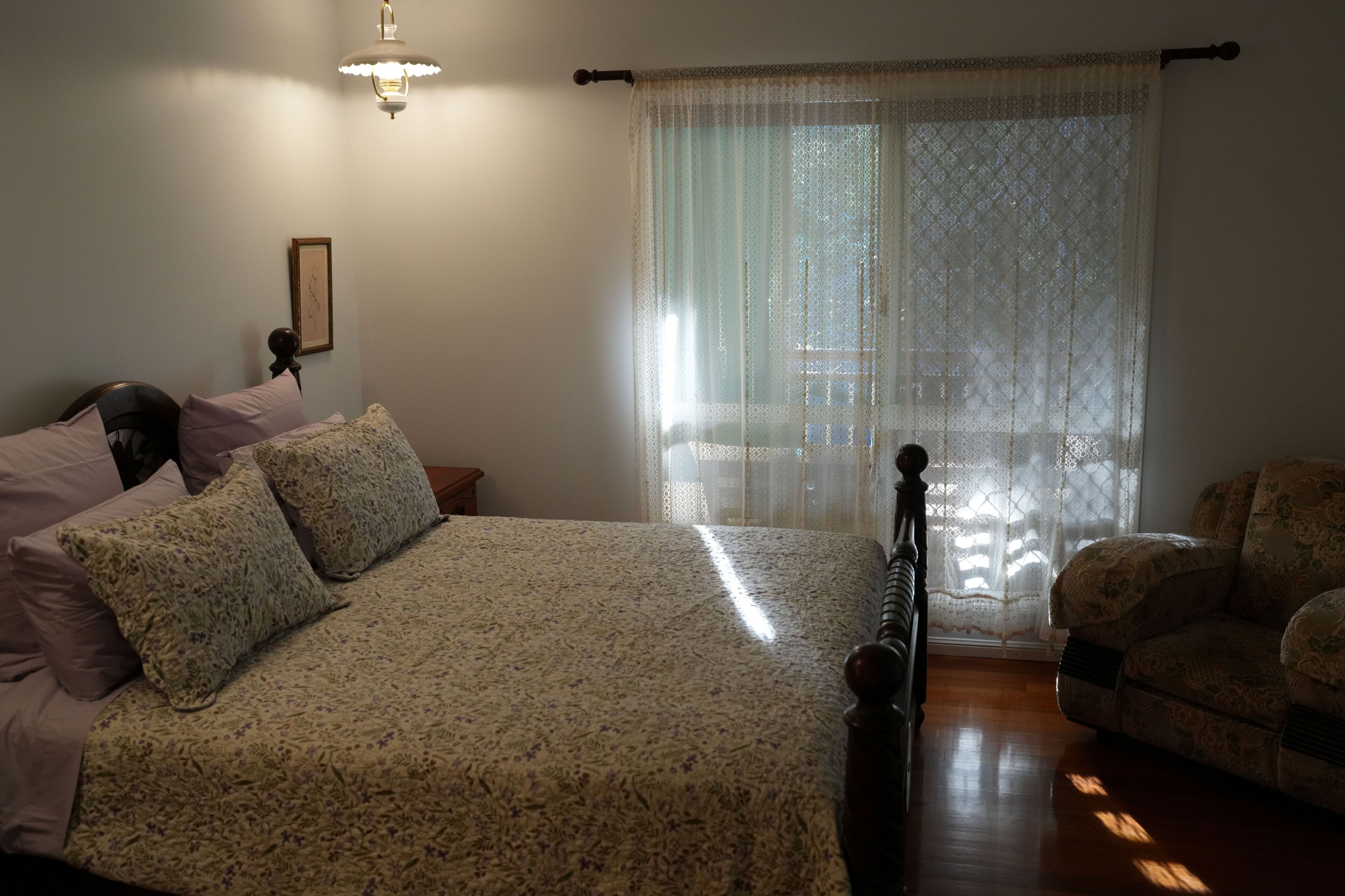 A bedroom with a bed, a chair, a nightstand, a lamp, and a window with lace curtains.