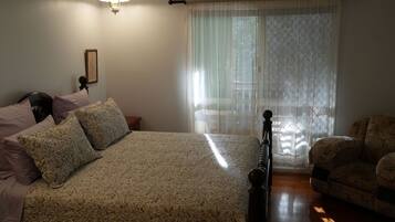 A bedroom with a bed, a chair, a nightstand, a lamp, and a window with lace curtains.