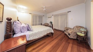 Un dormitorio con una cama y mesita de noche de madera, una silla, un ventilador de techo y una ventana con cortinas.