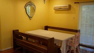 A room with a wooden bed, a small table, a chair, a mirror, an air conditioning unit, and a window with a curtain.