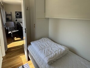 Ein Schlafzimmer mit einem Einzelbett, einem Sessel, einem Regal mit Büchern und einer Tür zu einem anderen Raum.