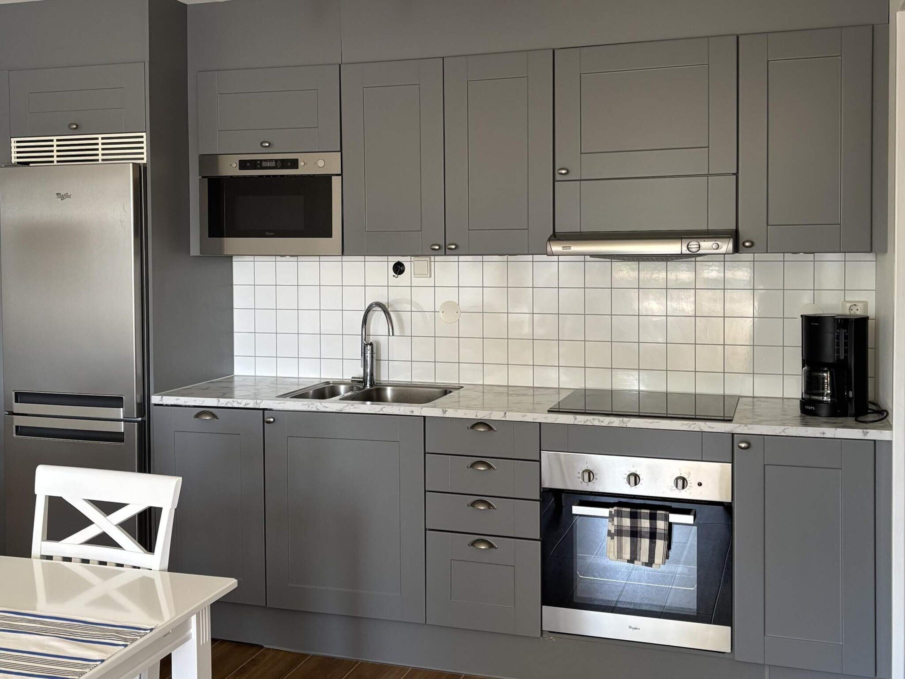 A modern kitchen with grey cabinetry, a stainless steel refrigerator, a microwave, a coffee maker, and a built-in oven.