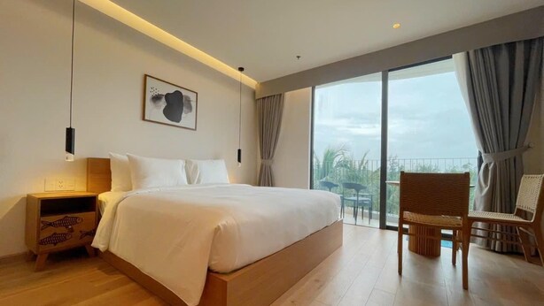 Un dormitorio moderno con una cama grande, una mesita de noche, una silla y vistas al exterior a través de un ventanal amplio.