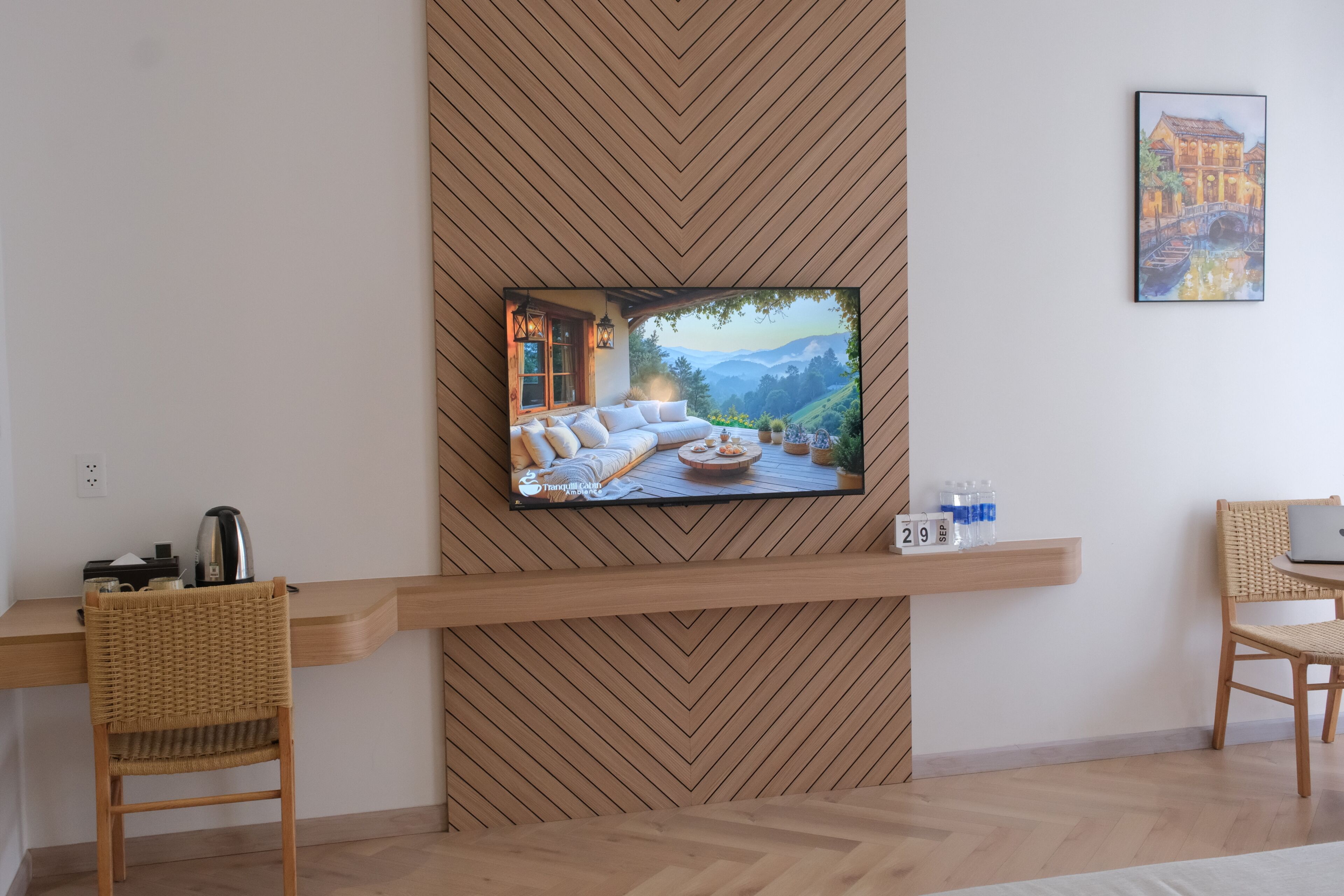 A modern room with a flat-screen TV mounted on a wooden wall, a wicker chair, a laptop on a small table, and a framed painting.