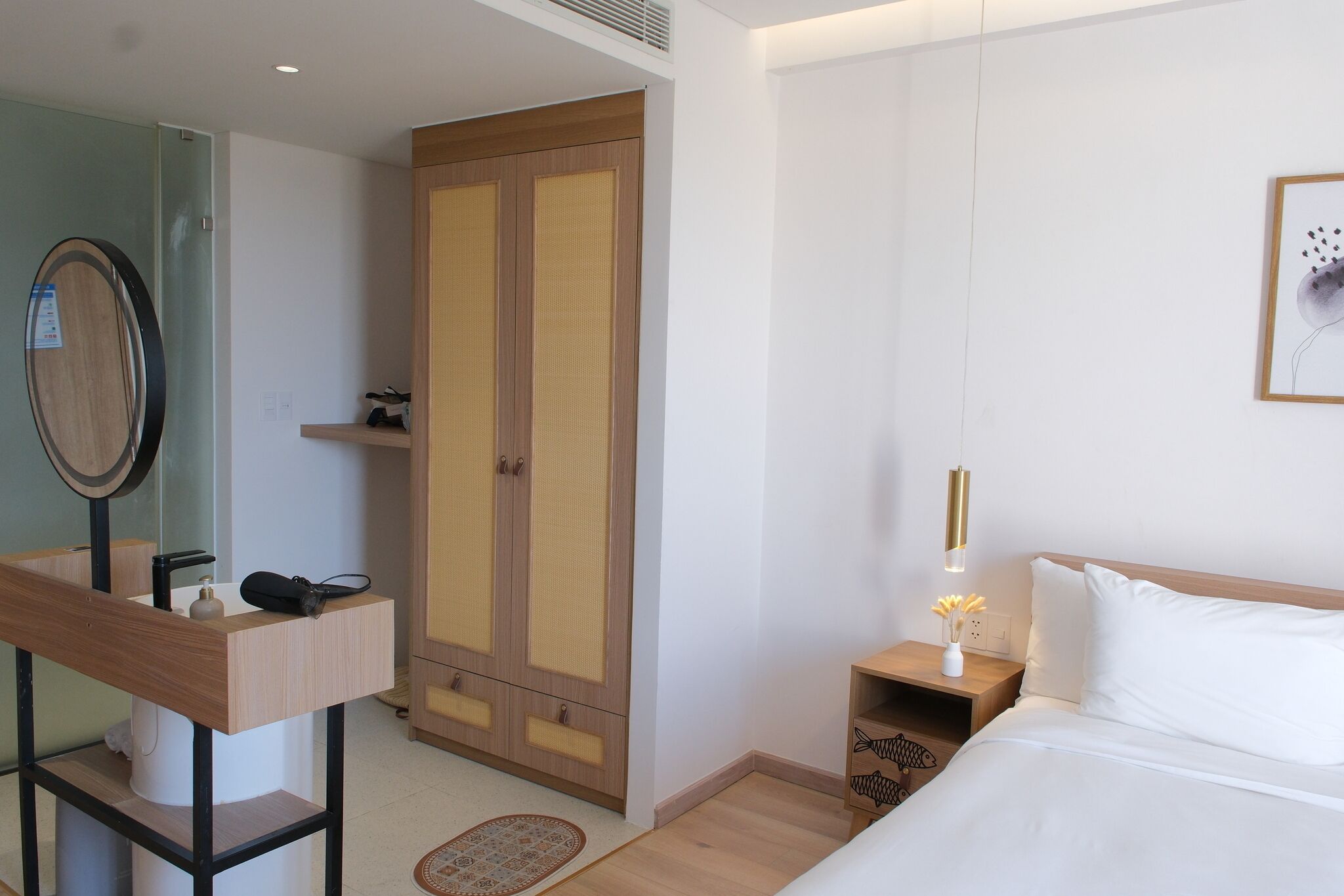 A hotel room with a wooden wardrobe, a bedside table with a lamp, a bed, and a mirror.