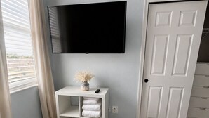 Un dormitorio con una cama, un televisor montado en la pared, una mesita de noche blanca con toallas y una puerta.