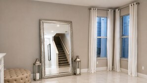 Una habitación amplia con un espejo grande, un otomano tapizado, una escalera y dos ventanas con cortinas.
