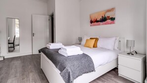 Ein ordentlich bezogenes Bett mit weißen Laken und einer grauen Decke, ein Nachttisch mit Lampe sowie ein Bild an der Wand.