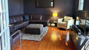 Un salon moderne avec un mur de couleur sombre, un canapé d’angle, un fauteuil blanc, une petite table ronde avec une lampe et un grand miroir.