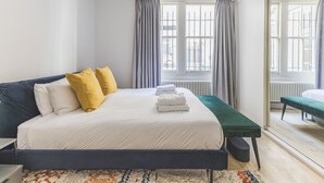 A bedroom with a bed, a bedside table, a bench, and a view of the outside through large windows. - Notting Hill Patio Retreat (London)