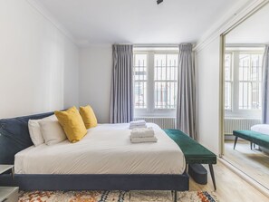 A bedroom with a bed, a bedside table, a bench, and a view of the outside through large windows. - Notting Hill Patio Retreat (London)