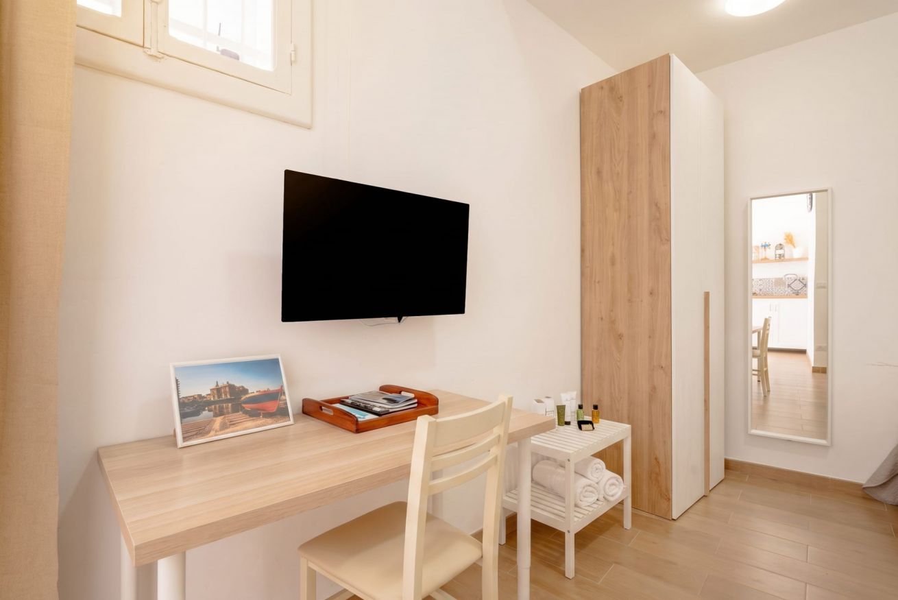 A modern room with a flat-screen TV mounted on the wall, a wooden desk with a chair, a small white shelf with toiletries, and a framed picture on the desk.