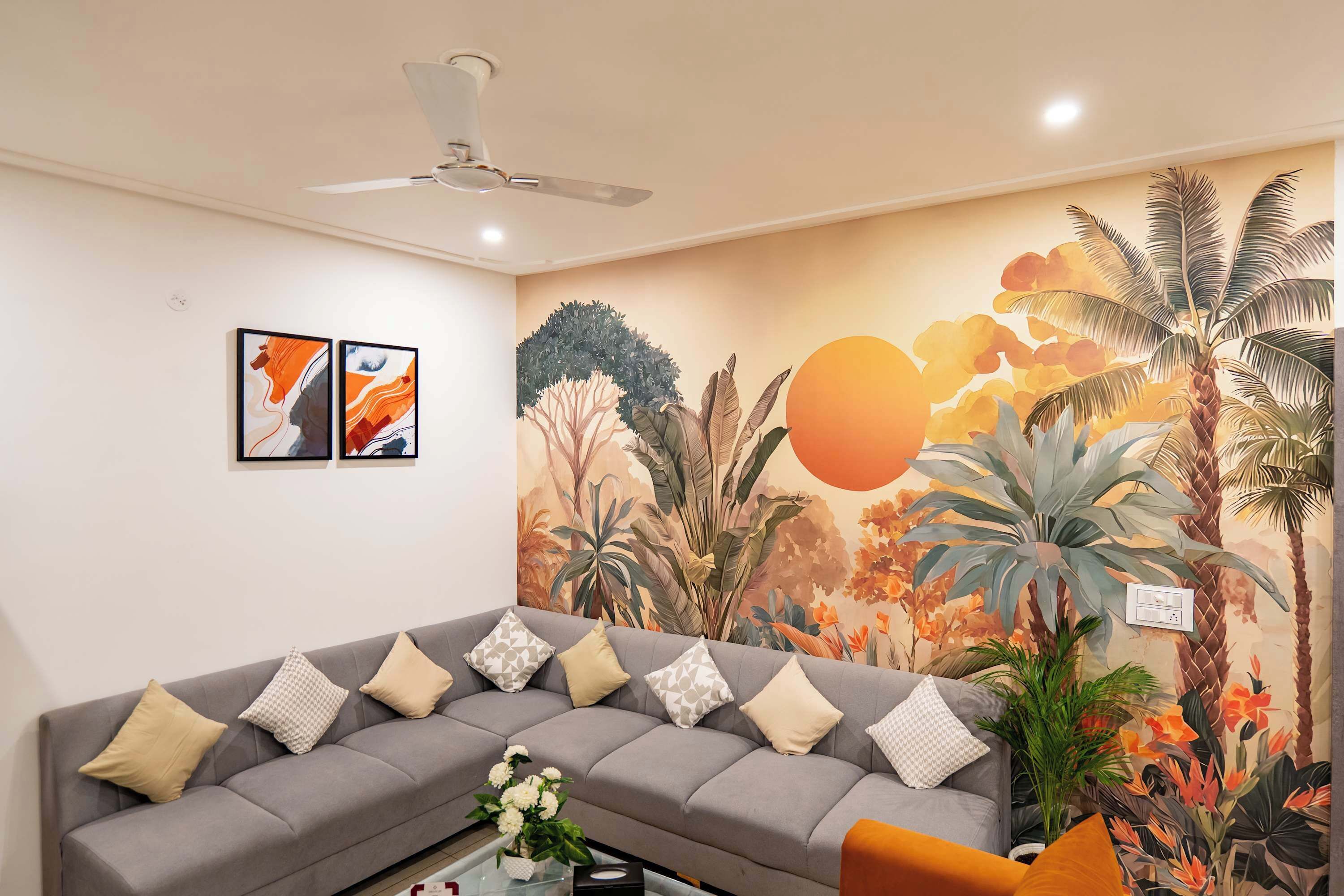 Una sala de estar con un sofá gris, un ventilador de techo y un mural en la pared con plantas tropicales.