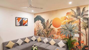 Una sala de estar con un sofá gris, un ventilador de techo y un mural en la pared con plantas tropicales.