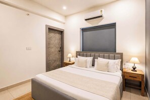 A hotel room with a bed, bedside tables, lamps, an air conditioning unit, and a door.