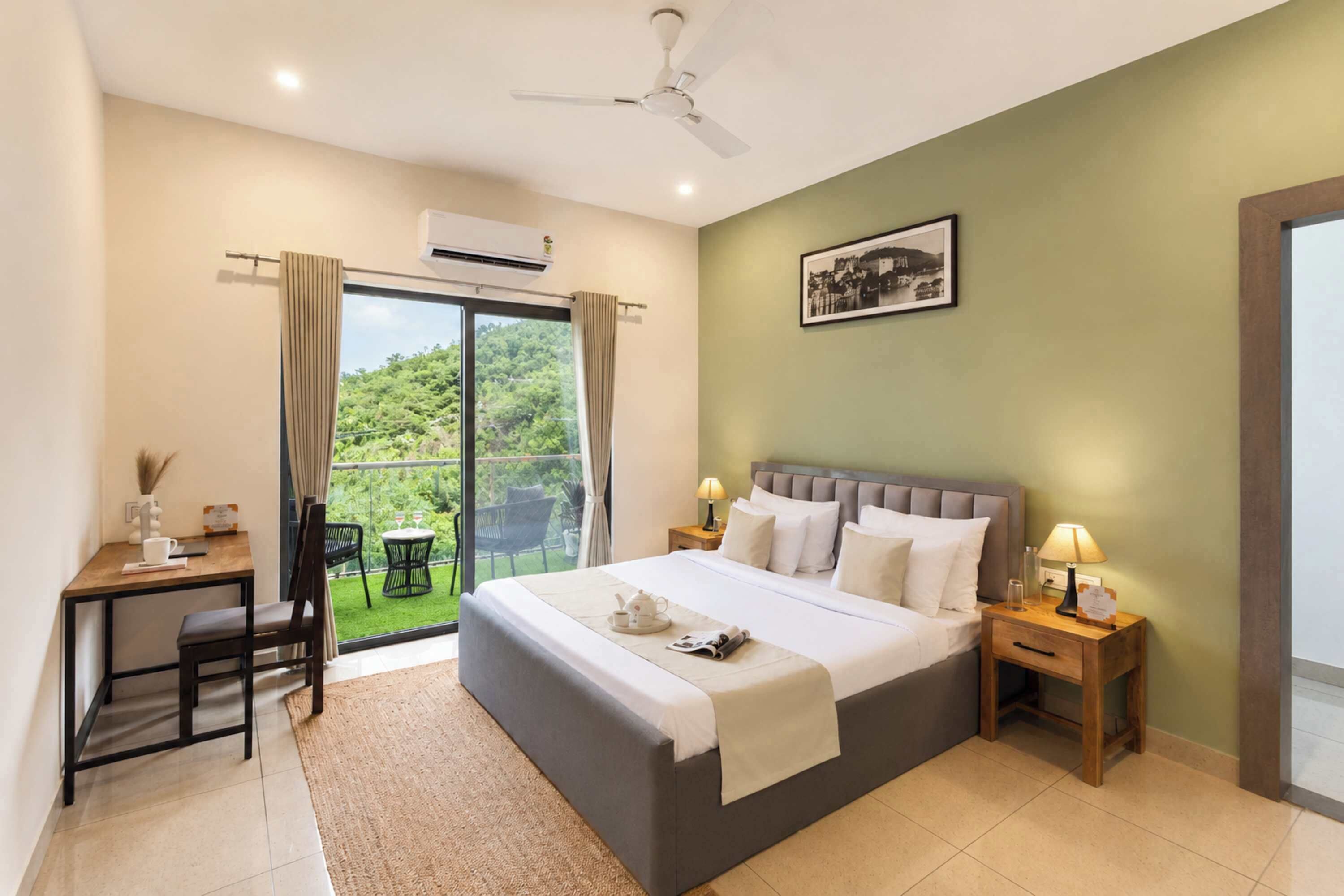 A hotel room with a bed, bedside lamps, a desk with a chair, and a view of greenery through a sliding door.