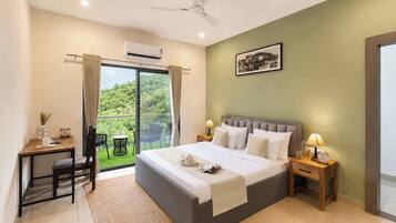 A hotel room with a bed, bedside lamps, a desk with a chair, and a view of greenery through a sliding door.