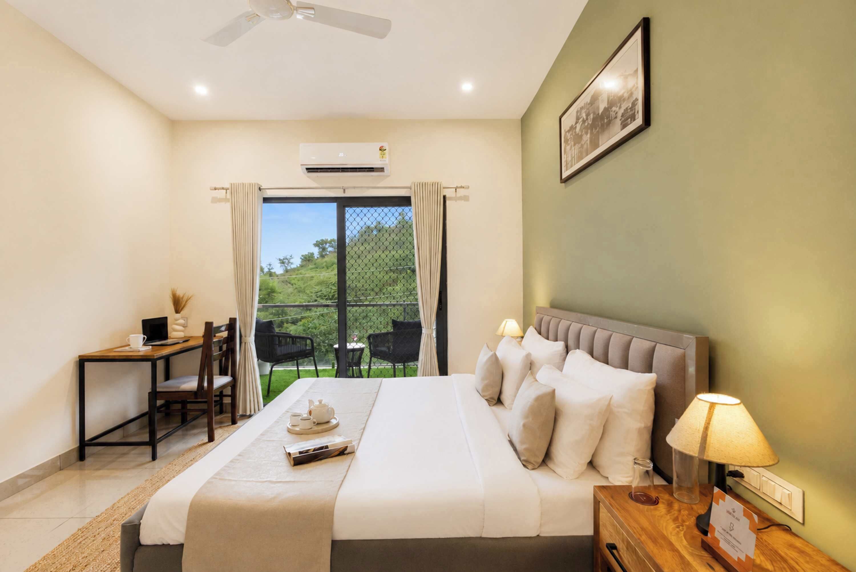 A hotel room with a bed, a desk, a chair, a table, a lamp, and a view of greenery outside.