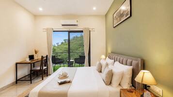 A hotel room with a bed, a desk, a chair, a table, a lamp, and a view of greenery outside.