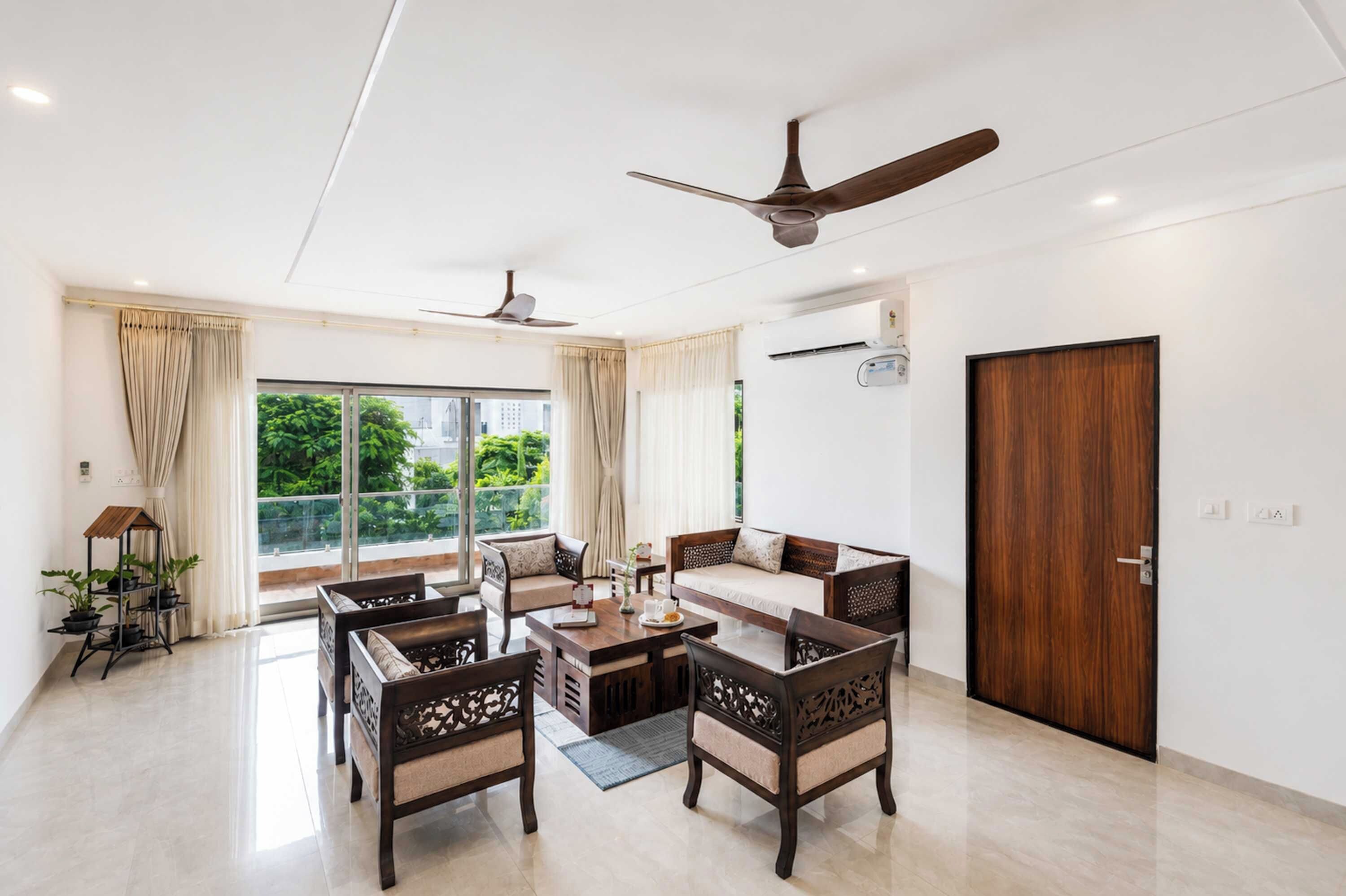 A spacious living room with a high ceiling, ceiling fans, a large window with curtains, a wooden door, and a dining area with a table and chairs.