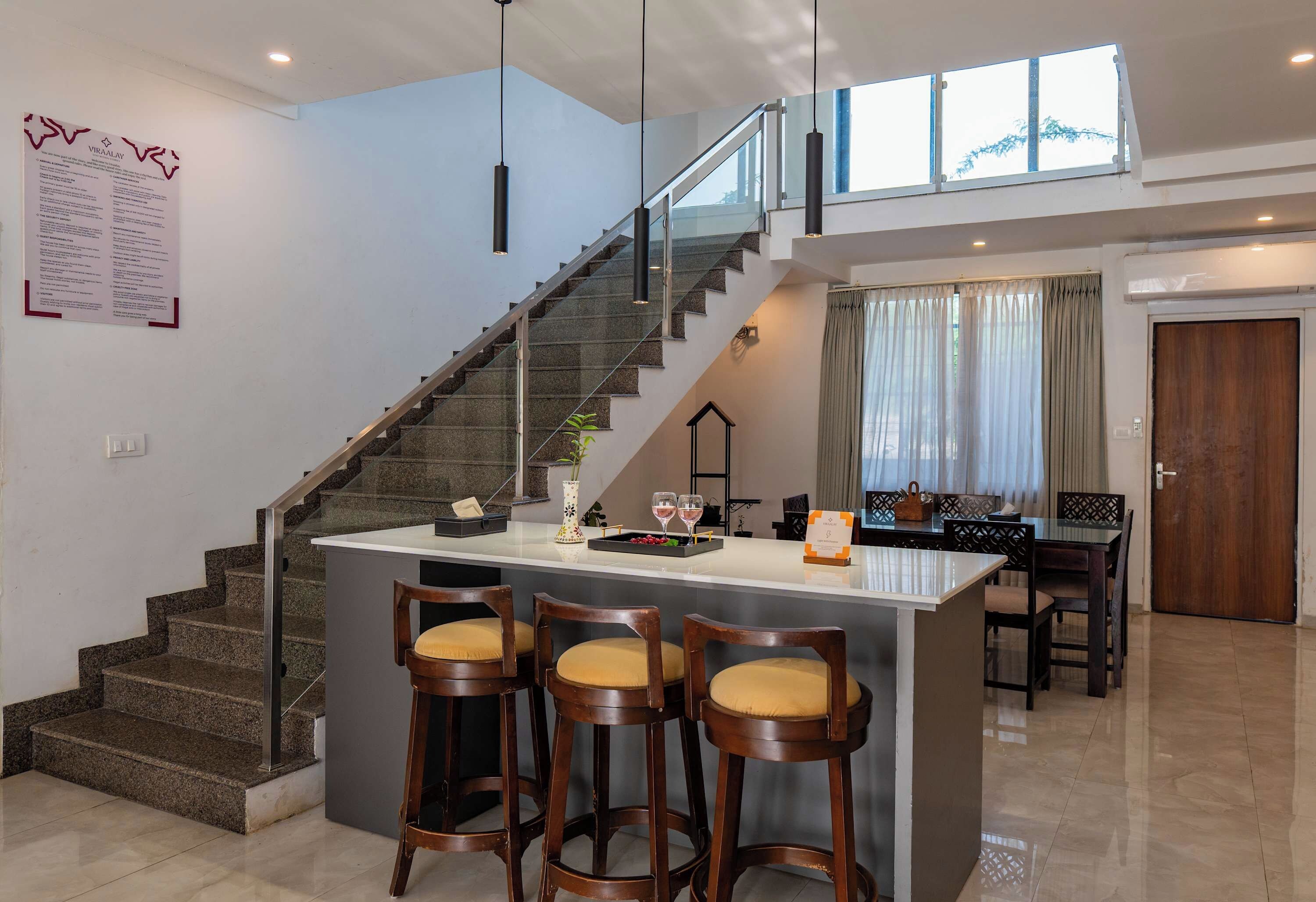 A modern kitchen with a central island, bar stools, a staircase, and a dining area with a view.