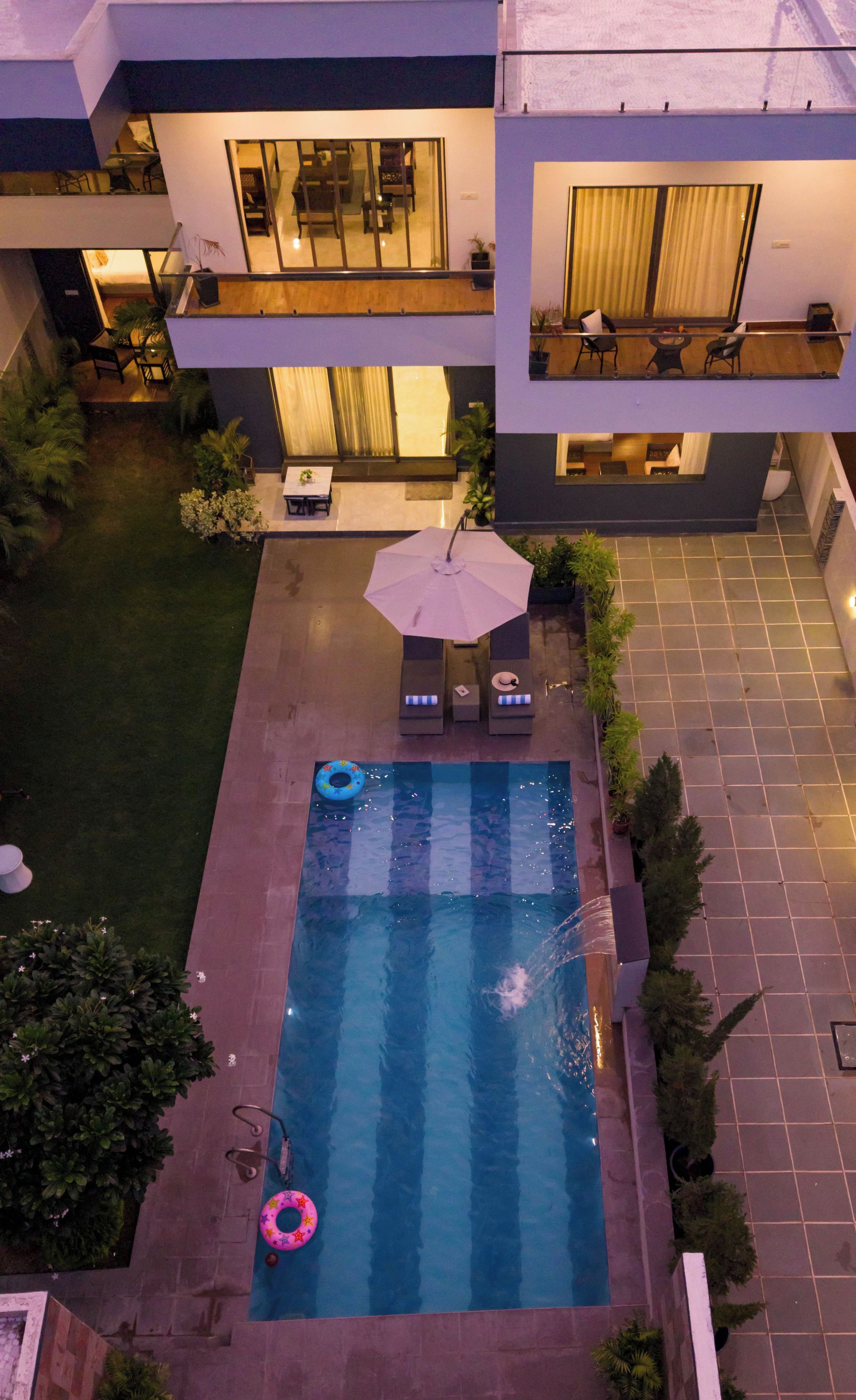A rooftop pool with lounge chairs and an umbrella, surrounded by a garden and a building with a balcony.