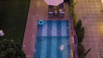 A rooftop pool with lounge chairs and an umbrella, surrounded by a garden and a building with a balcony.