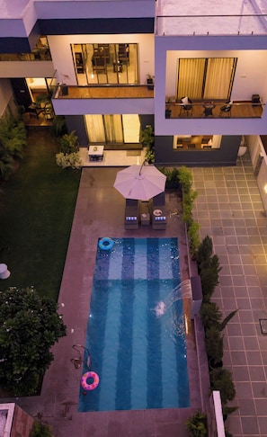 Una piscina en la azotea con sillones de descanso y una sombrilla, rodeada de un jardín y un edificio con balcón.