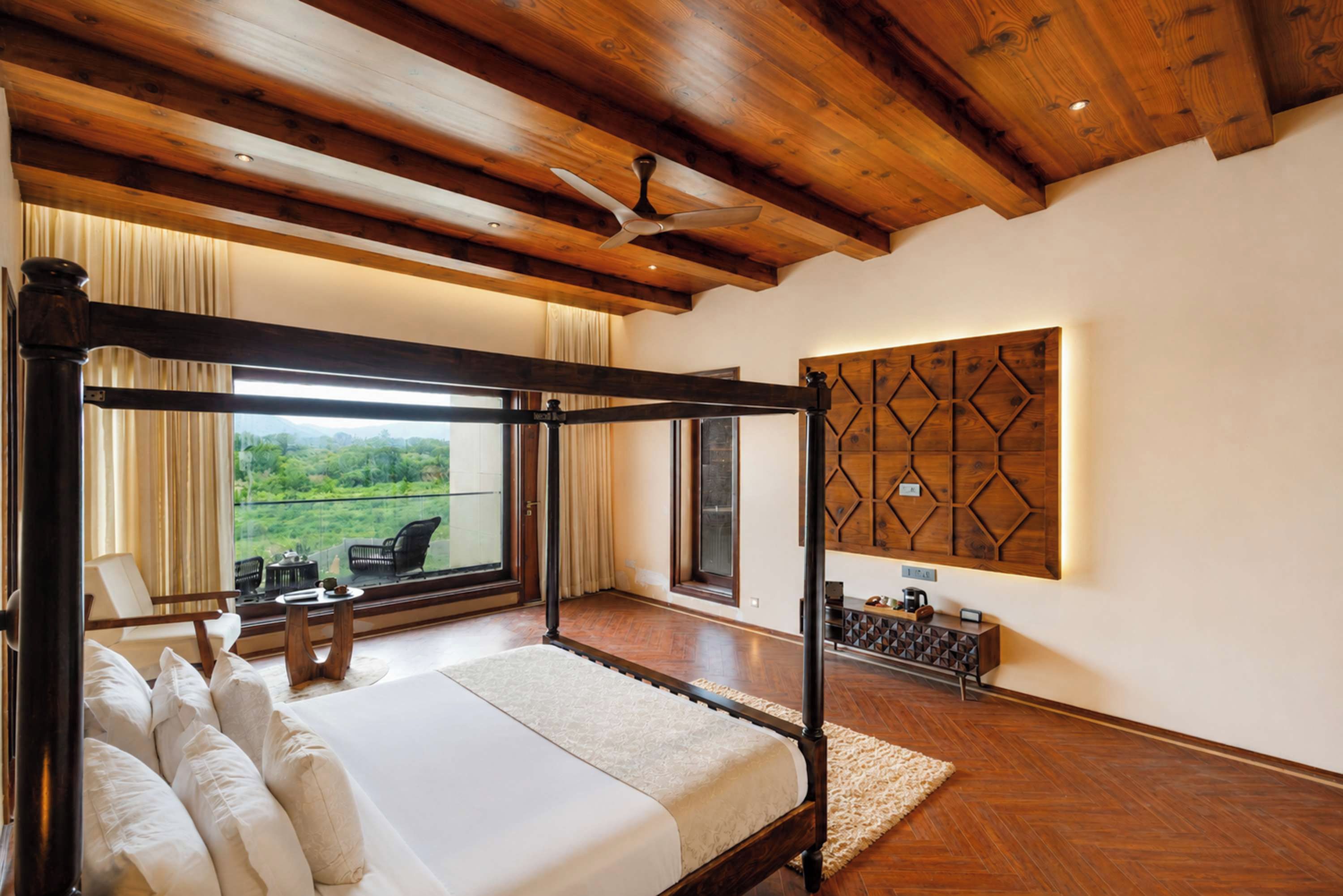 A four-poster bed with a view of greenery outside, a ceiling fan, and a decorative wall panel.