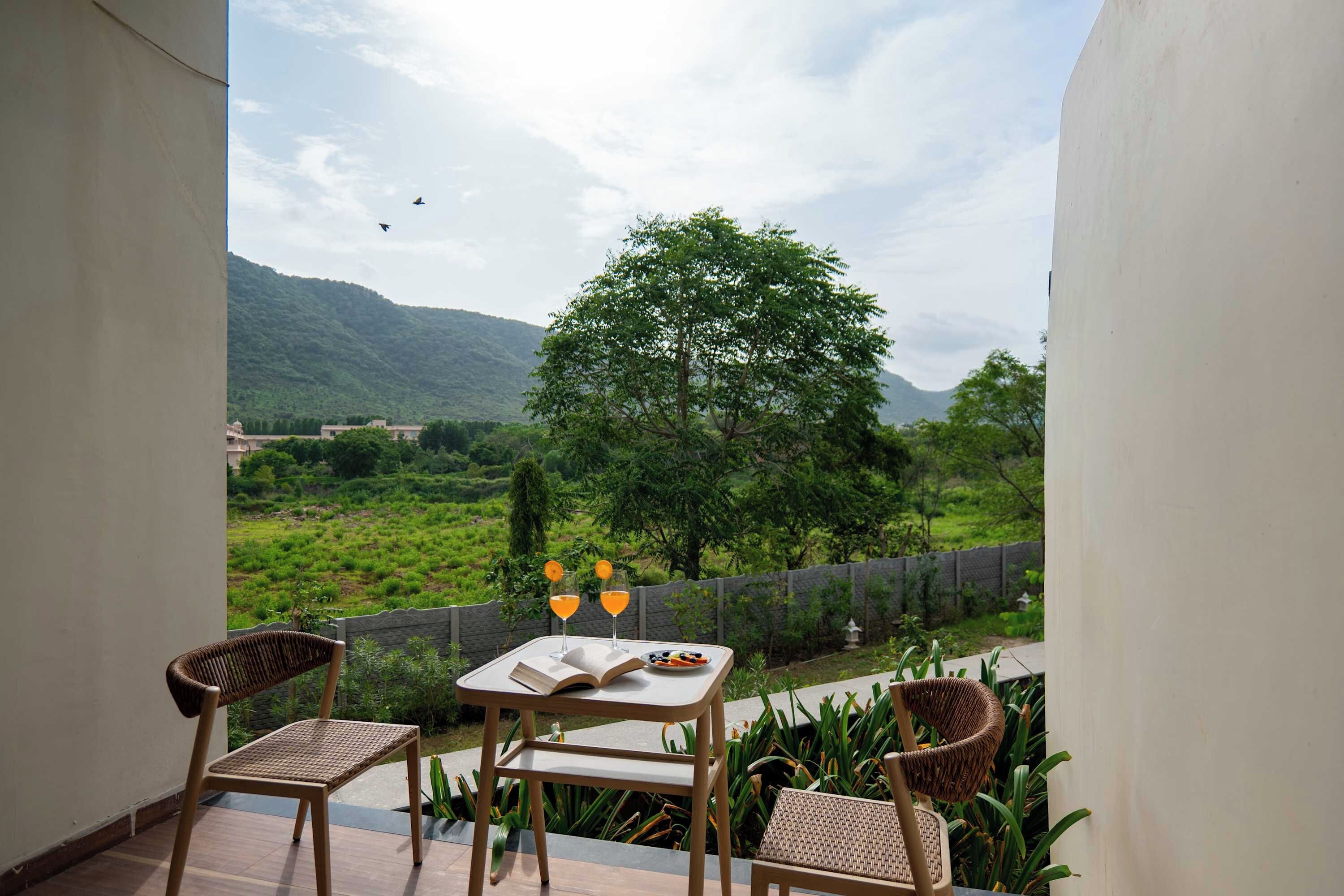 A balcony with two wicker chairs, a table with a book and two glasses of orange juice, and a view of a green landscape with a tree and mountains in the distance.