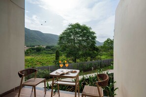 Een balkon met twee rieten stoelen, een tafel met een boek en twee glazen sinaasappelsap, en uitzicht op een groen landschap met een boom en bergen in de verte.