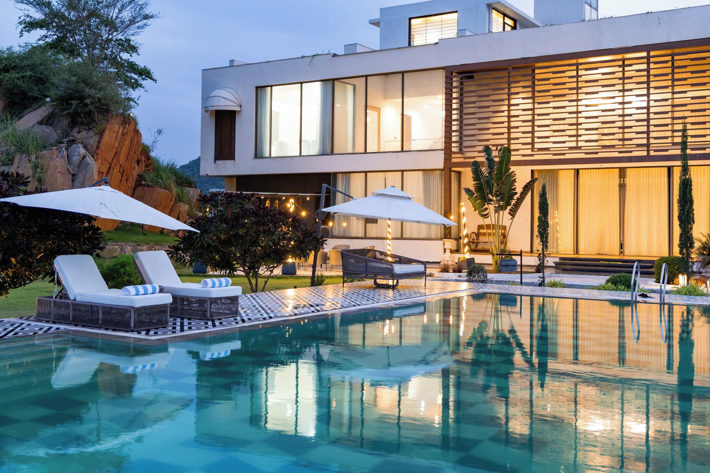 A modern pool area with lounge chairs and umbrellas, surrounded by a landscaped garden and a contemporary building.
