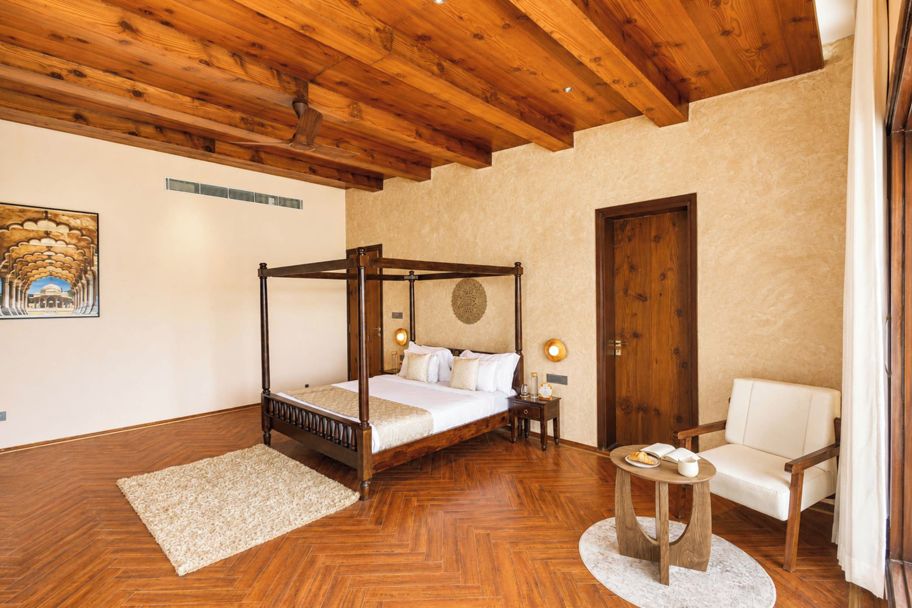 Ein Bett mit Baldachin und Holzfußteil, ein runder Tisch mit weißer Tischdecke sowie ein weißer Sessel in einem Raum mit Holzboden und hohen Decken.