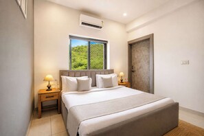 A hotel room with a large bed, two bedside tables with lamps, a window with a view of greenery, and an air conditioning unit.
