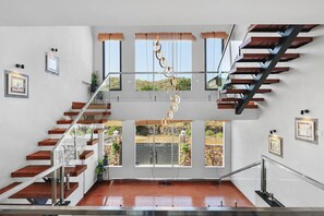 A modern interior with a glass staircase, wooden railings, and large windows offering views of the outdoors.