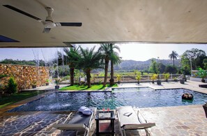 A pool area with a stone floor, lounge chairs, a table with glasses, and a view of a landscaped garden with palm trees and a stone wall.