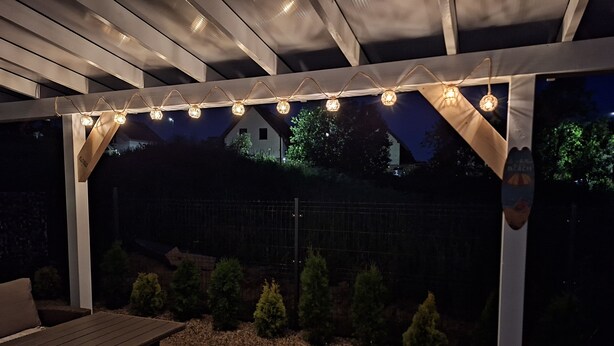 Ein überdachter Patio-Bereich mit Lichterketten, einer Holzbank und einem Garten mit kleinen Bäumen und Kieselsteinen.