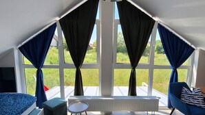 Ein modernes Zimmer mit einem Bett, einem Sessel, einem kleinen Tisch und einem großen Fenster mit Vorhängen.