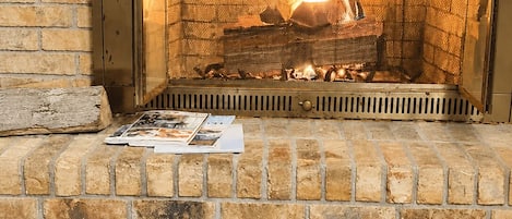A fireplace with a brick surround, a magazine on the mantel, and a wooden log fire.