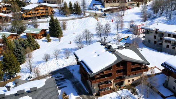 Ein schneebedecktes Chalet mit Skipiste im Hintergrund, Holzfassade und Parkplatz.