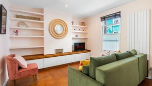 Ein modernes Wohnzimmer mit einem grünen Sofa, einem rosafarbenen Sessel, einem Holztisch, einem Netflix-Fernseher und einem Fenster mit Jalousien.