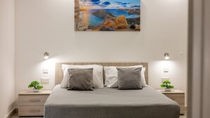Ein Schlafzimmer mit einem Bett, zwei Nachttischen, einem kleinen Tisch und einem Gemälde einer Küstenlandschaft. Auf jedem Nachttisch steht ein kleiner Blumentopf.