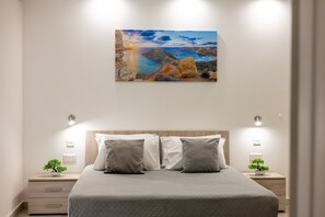 Ein Schlafzimmer mit einem Bett, zwei Nachttischen, einem kleinen Tisch und einem Gemälde einer Küstenlandschaft. Auf jedem Nachttisch steht ein kleiner Blumentopf.
